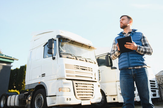 Handsome Caucasian man wearing red cap standing next to trucks while using tablet device. Hardworking driver typing message or text for delivery order. Logistics company concept