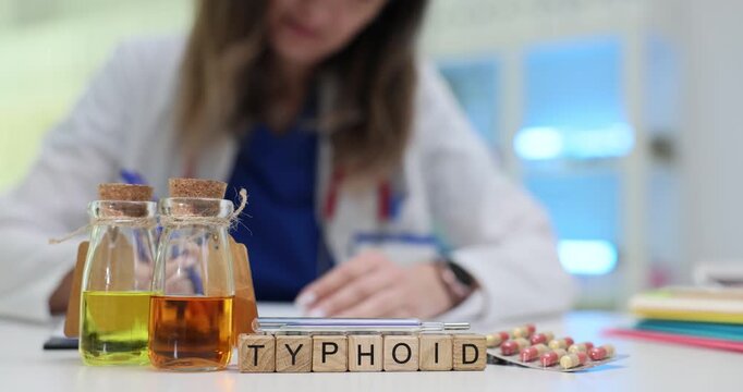 Wooden blocks spell Typhoid put near bottles with colored liquid on table. Light enters room creating medical atmosphere with capsules and test tubes