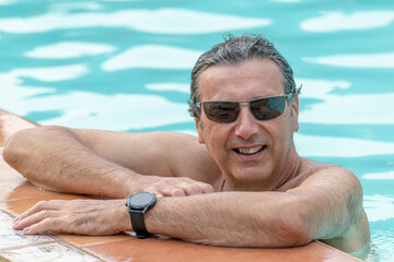A handsome wet man is leaning with his arms on the edge of the pool, with his body in the water