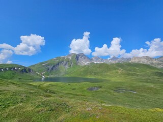La Thuile, Val D'Aosta, July 02 2025: Panoramic view to the alps, beautiful green landscape reflected in a small natural lake. Beautiful blue sky with some clouds. Mountains with a little snow. Aerial