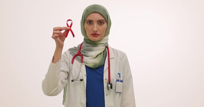 Woman doctor in hijab holds red ribbon symbol of AIDS and condoms against white wall. Physician raises awareness about safe sex practices. Slow motion