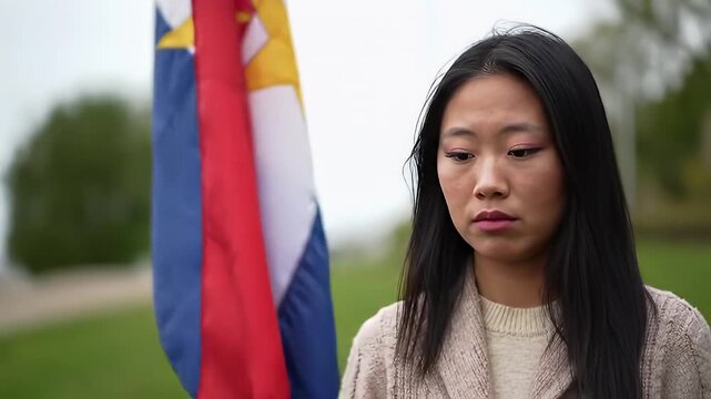 A young woman stands somberly next to a blurry flag, with grass and sky in background