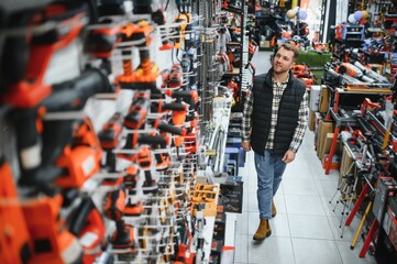 Obraz premium Portrait of young bearded man buying modern electric drill while standing in tool. Hardware construction site objects concept