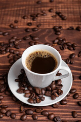 A cup of dark roasted coffee on a white saucer, surrounded by coffee beans on a brown wooden surface