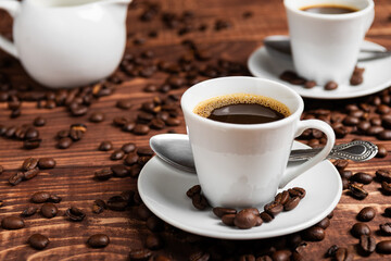 A cup of dark roasted coffee on a white saucer, surrounded by coffee beans on a brown wooden surface
