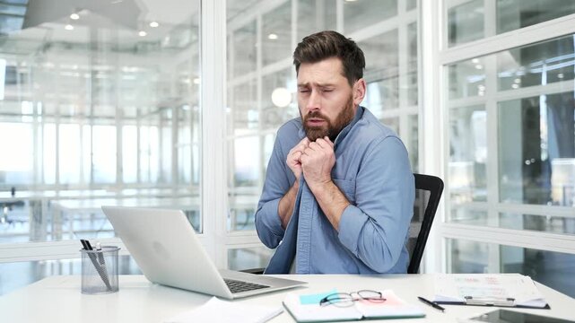 Businessman froze while working on laptop sitting at workplace in a modern business office. Upset male cold indoors. Unhappy worker feels discomfort due to low temperature. Problems with heating