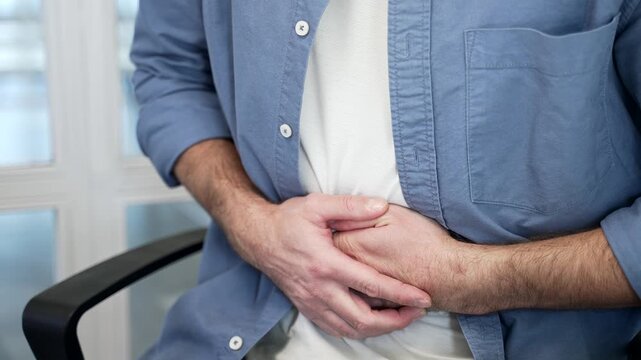 Close up. Businessman in blue shirt feels stomach pain sitting at workplace in business office. Male hands holding massaging stomach. Sick man worker suffers from gastritis, poisoning or bloating