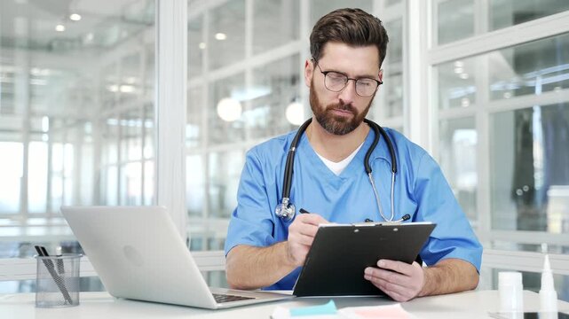 Busy doctor in blue coat working at workplace writing down data using laptop sitting at desk in hospital office. Medical worker physician doing paperwork, filling out form or writing out prescription