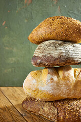 Stack of assorted rustic bread loaves arranged on a wooden table with a textured green wall background, showcasing artisan bakery style, natural food photography, and organic homemade bread concept
