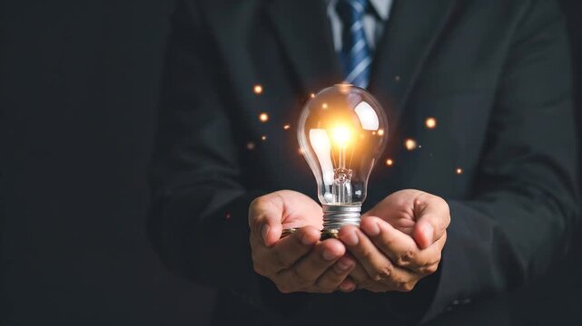 A person in a suit cradles a glowing lightbulb, emanating sparks. The background is dark. A visual representation of an idea
