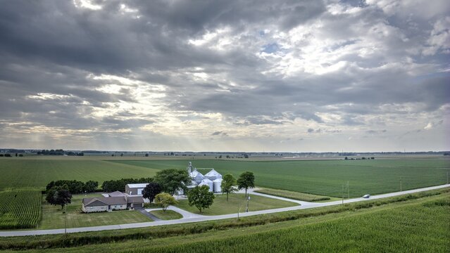 Aerial view of a farm with silver silos gleaming under the dramatic sky, contrasting with the verdant fields stretching to the horizon, Monroeville, Indiana, United States.