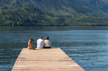 Couple with dog sitting on the wooden dock by the beautiful lake with forested mountains