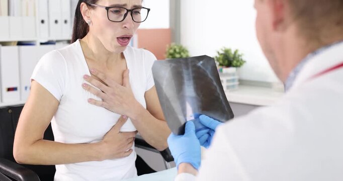 Woman coughs holding chest while doctor inspects lung X-ray during appointment. Posture indicates breathing problem creating urgency in consultation
