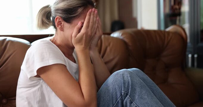 Woman sits on couch crying and hiding face with both hands in emotional pain. Sadness builds overwhelming body creating fragile and broken moment