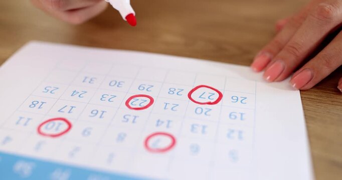 Marker highlights day on printed calendar creating visible circle around number. Woman hand presses tip firmly making bright red mark on sheet