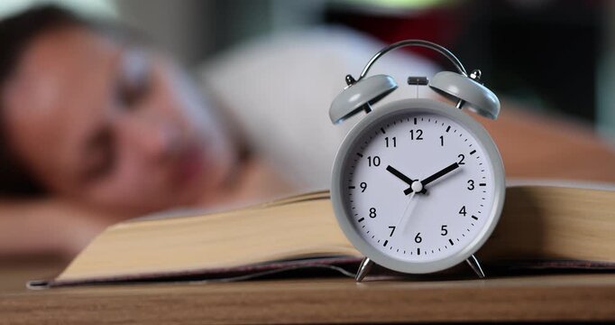Alarm clock stands near open book resting near sleepy student creating wake-up moment. Timepiece shows late hour disturbing learner during quiet nap