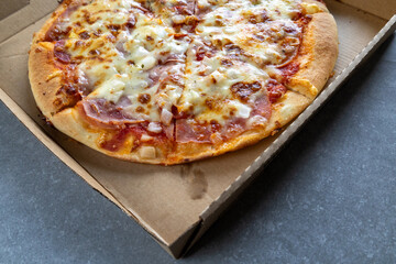 Side view of a freshly baked pizza topped with melted cheese, ham, onions, and tomato sauce inside a cardboard delivery box. 