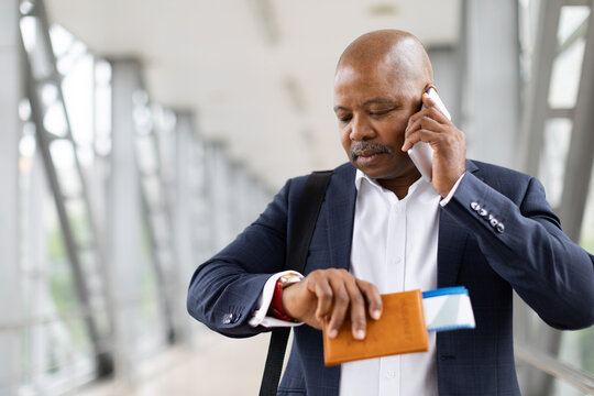 Focused African American businessman checking watch while talking on smartphone and holding passport at airport terminal. Business travel and punctuality concept, closeup