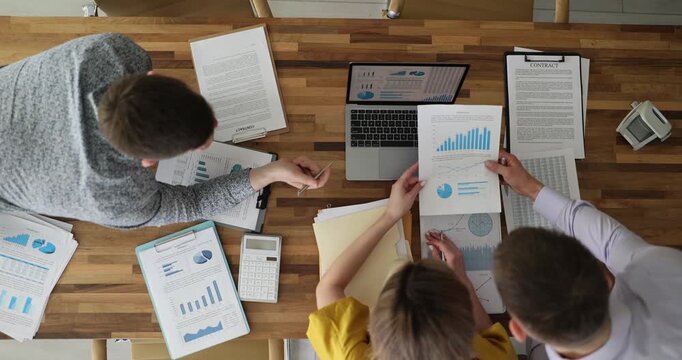 Coworkers team gathers around laptop and papers during busy work session. Hands point and arms move rapidly sorting through charts and data on table