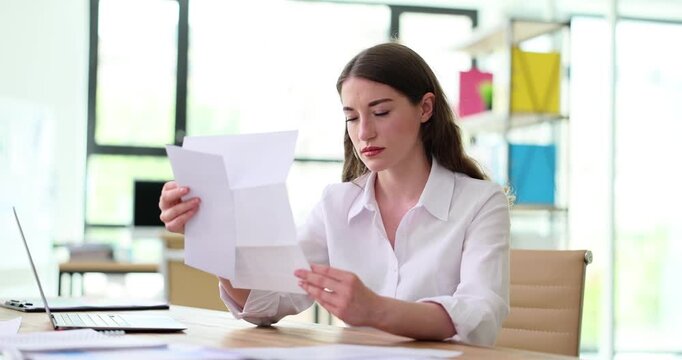 Woman sits at desk and intently reads document got by mail held in hands. Concept of serious concentration and focus on important paperwork in office