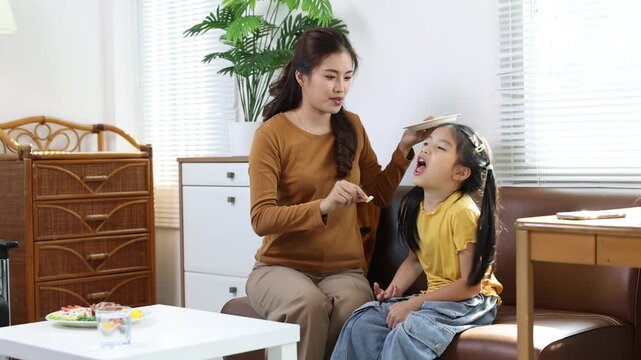 Mother feeding picky daughter at home. Mom patiently tries to snack her reluctant young daughter, offering bites from a plate while sitting on a comfortable sofa in their living room.
