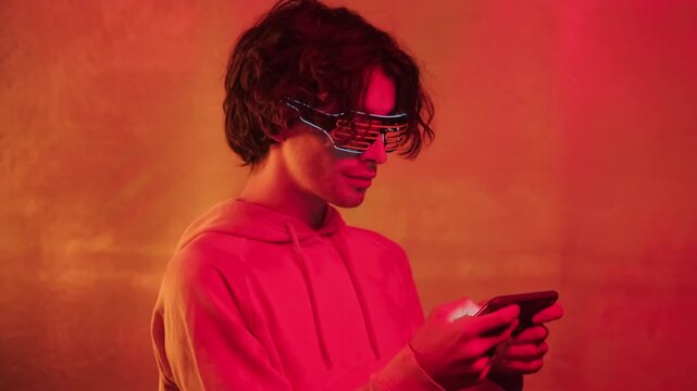 Medium shot of stylish young brunette man in neon shutter glasses using his smartphone standing on bright orange background