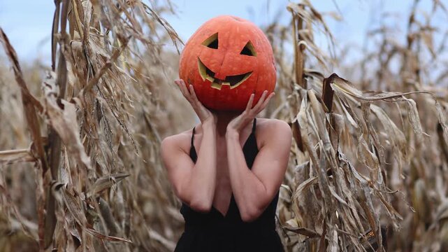 Woman in spooky carved Halloween pumpkin head and long dark dress stands at autumn dry corn field, camera moves. Jack Lantern. Trick o treat. Creepy carnival costume for celebration mystical party