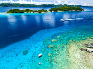 Naklejka premium 大型フェリーのざまみ号が入港しようとしている。 座間味島や安慶名敷島が見える。 ボートシュノーケリングに出港して、スキンダイビングをしている。 許可を得てドローンで空中撮影をしている。 巨大なサンゴ礁と熱帯魚の群れ。 沖縄県島尻郡慶良間諸島座間味島嘉比島-2025/6/27 