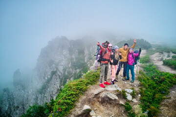Group of four hikers, equipped with backpacks and trekking poles, cheerfully pose on misty mountain ridge. Rocky cliffs, lush greenery around create dramatic backdrop, while fog adds mystical touch.