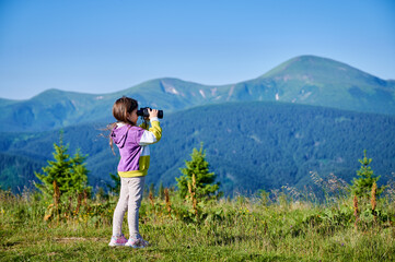 Naklejka premium Young girl stands in grassy meadow, looking through binoculars, wearing yellow and purple sweatshirt. Behind her, rolling green hills and mountains stretch into distance under clear blue sky.