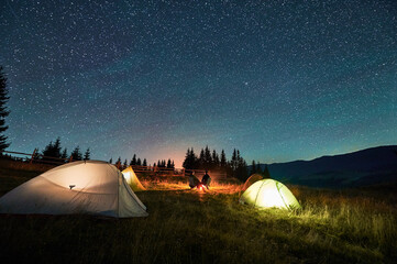 Silhouette of couple hikers resting by campfire under star-studded sky, near illuminated tents at campsite. Night camping in mountains, night sky create magical and peaceful atmosphere.