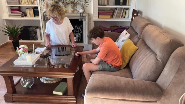 Mother and her 12-year-old son playing dominoes on a glass coffee table in the living room, smiling and enjoying a playful and cozy moment together during an indoor family game