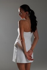 Elegant young woman in a white strapless dress and high heels posing in studio. Fashion model with long hair and red lips, sitting gracefully on a white background. Beauty, fashion and portrait