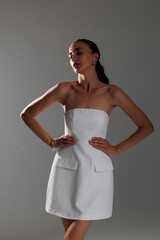 Elegant young woman in a white strapless dress and high heels posing in studio. Fashion model with long hair and red lips, sitting gracefully on a white background. Beauty, fashion and portrait