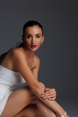 Elegant young woman in a white strapless dress and high heels posing in studio. Fashion model with long hair and red lips, sitting gracefully on a white background. Beauty, fashion and portrait