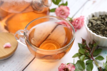 Tasty rose tea in glass cup, flowers and leaves on white wooden table, closeup