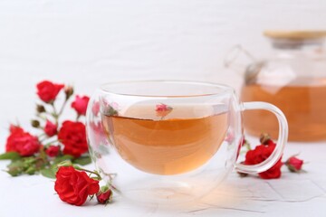 Aromatic tea in glass cup, roses and green leaves on white table, closeup