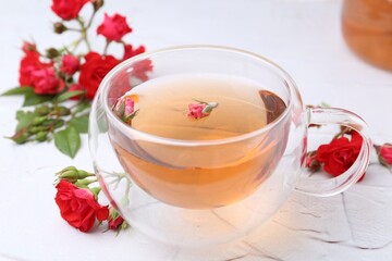 Aromatic tea in glass cup, roses and green leaves on white table, closeup