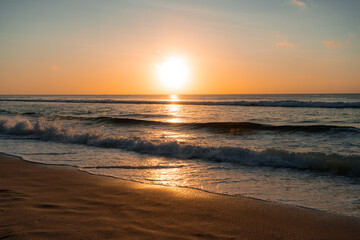Horizontal photo of golden sunset over the ocean with gentle waves crashing on the sandy beach, reflecting warm orange light on the water and shore