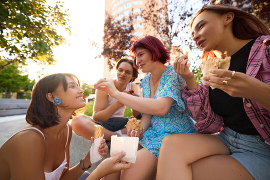 Young woman feeding friend with french fry during outdoor snack with group. Concept of emotional storytelling about friendship, youth lifestyle, social media blogging, street food promotion.