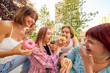 Group of young friends laughing and sharing donuts outdoors in sunny urban park. Concept of friendship, joy, indulgence, street food, summer leisure, and positive emotions.