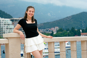 A young beautiful girl poses in summer on the resort's city promenade against the backdrop of the sea and mountains, dressed in a black top and a white short skirt