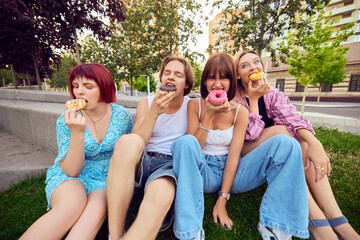 Happy friends sitting on grass and eating assorted colorful donuts together. Concept of outdoor leisure, joyful friendship, tasty treats, youth lifestyle, and sweet indulgence.