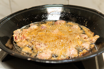 Close-up of raw pork meat being fried in hot oil inside a black pan, stirred with a metal spatula. Traditional Asian cooking process in a home kitchen showing authentic culinary preparation.