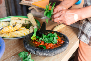 A woman prepares traditional Indonesian sambal using a stone mortar and pestle with fresh tomatoes and herbs. Authentic local cooking process that highlights Asian culinary culture and lifestyle.
