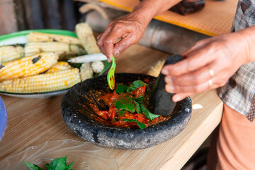 A woman prepares traditional Indonesian sambal using a stone mortar and pestle with fresh tomatoes and herbs. Authentic local cooking process that highlights Asian culinary culture and lifestyle.