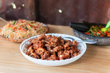 Close-up of crispy fried pork served in a white bowl on a wooden table, with stir-fried noodles and traditional chili sambal in the background. Authentic Asian cuisine and homemade food culture.