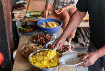 A group of people serving traditional Indonesian yellow rice with peanuts, fried noodles, and various side dishes during a communal meal or cultural gathering on a wooden table.