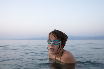 Naklejka premium Boy resting in swimming goggles, floating peacefully on calm lake waters during golden sunset, nature embracing swimmer's tranquil moment
