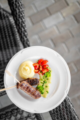 Grilled meat skewers with mashed potatoes, cherry tomatoes, and green herb sauce on a white plate, served on a woven surface with tiled background